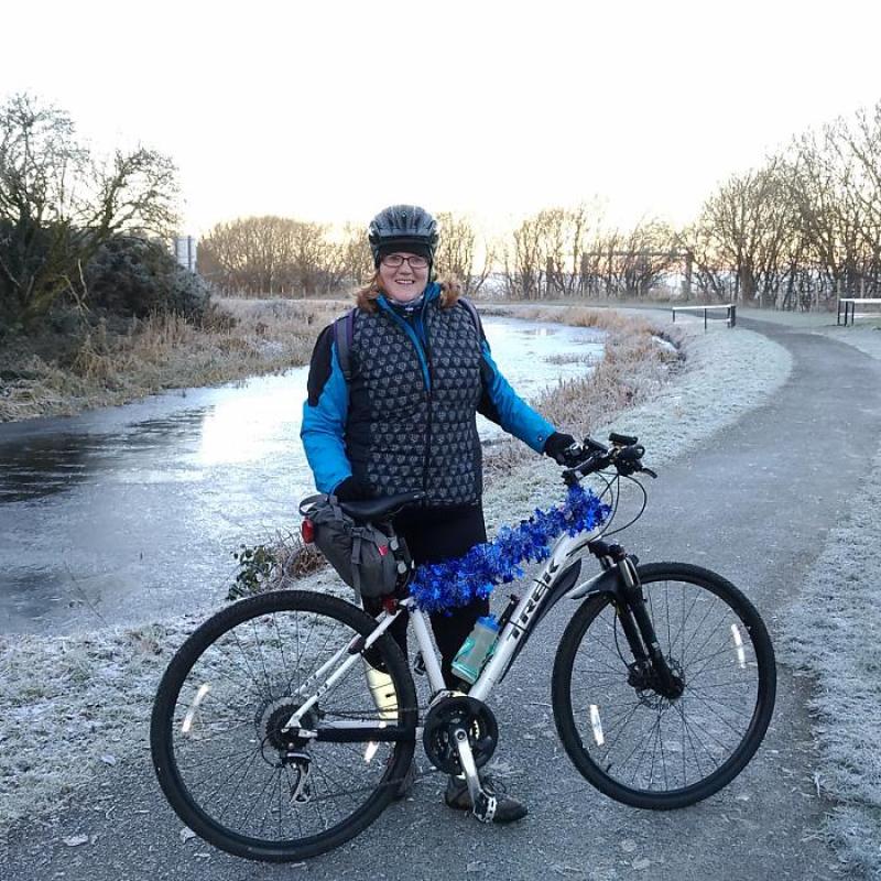 A woman is standing with a light grey Trek mountain bike. It’s winter. She is wearing a warm winter jacket and trousers. Her bike is decorated with blue tinsel. There is frost on the pavement, grass and trees and the stream behind her has ice on it