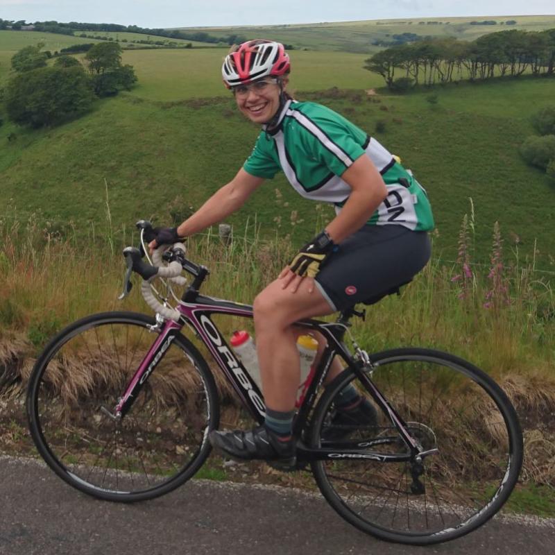 A woman is cycling on an Orbea road bike. She is wearing a green and white cycling jersey, black shorts, a red and white helmet and black and yellow fingerless gloves. She is smiling at the camera. In the background is some pretty impressive countryside