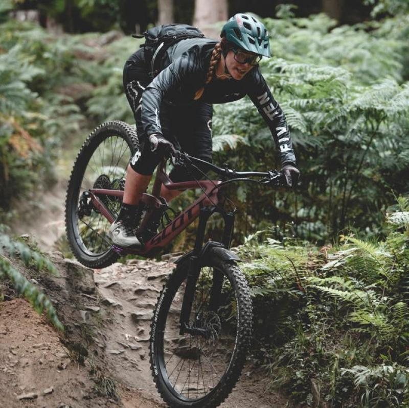 A woman is cycling downhill through a forest. She is riding a Scott mountain bike. She is wearing black three-quarter-length cycling leggings and a black jacket, as well as a green helmet. Credit: Roo Fowler
