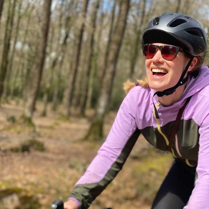 A woman in a lilac and forest green cycling jacket and silver helmet is cycling off road through a forest. It’s autumn and the trees are bare, there are leaves all over the forest floor