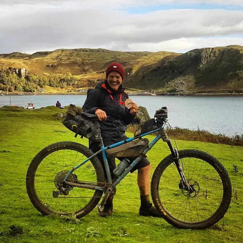 A woman is standing with a very muddy bike and holding a medal, having just completed an off-road triathlon on the Isle of Kerrera. She is smiling broadly
