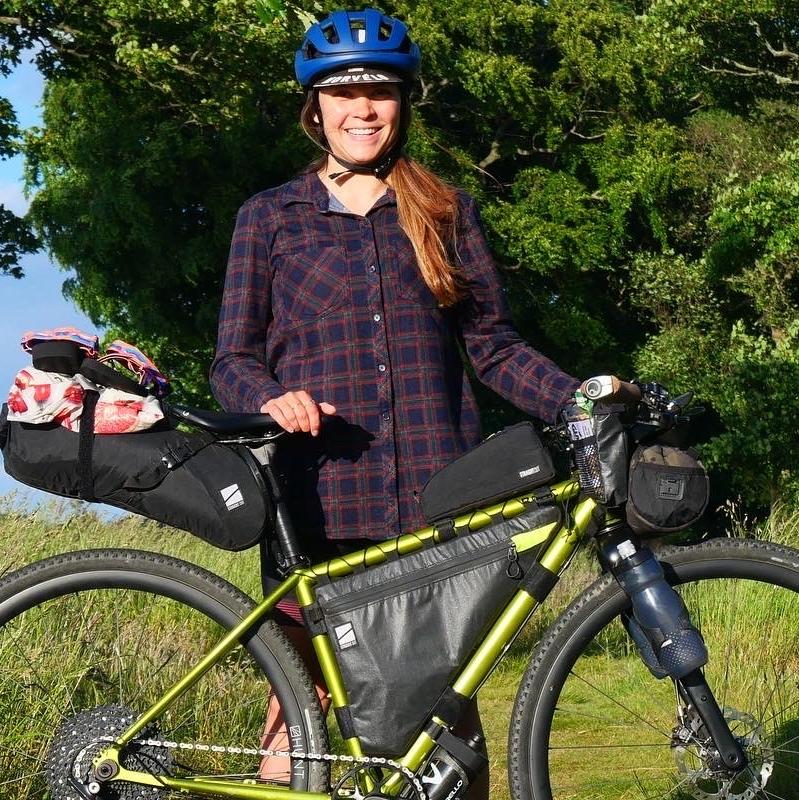 A woman is standing with a metallic green bike that is fully loaded with bags for bikepacking. She is wearing a blue cycling helmet and a check shirt. She is smiling at the camera