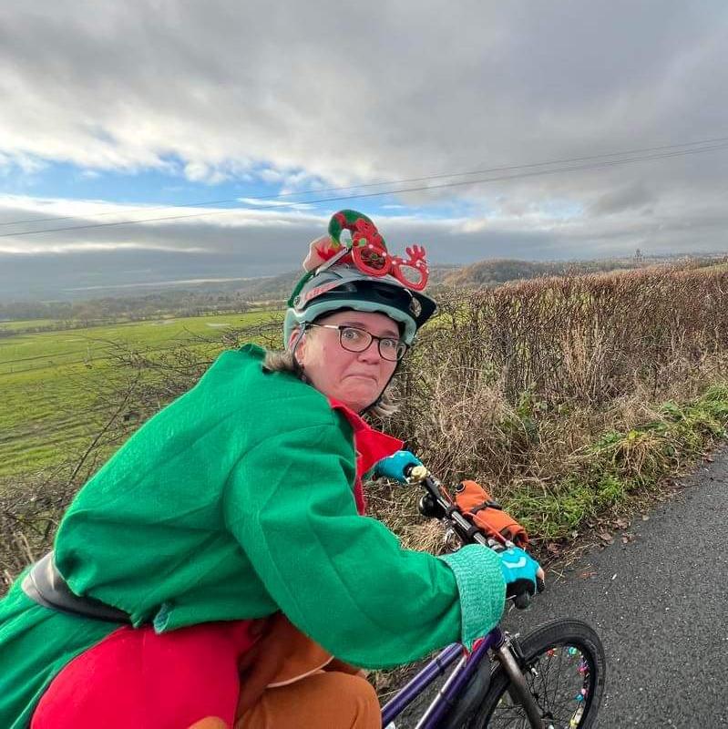 A woman is riding a bike while wearing a Christmas elf outfit. She is looking back at the camera and pulling a funny face