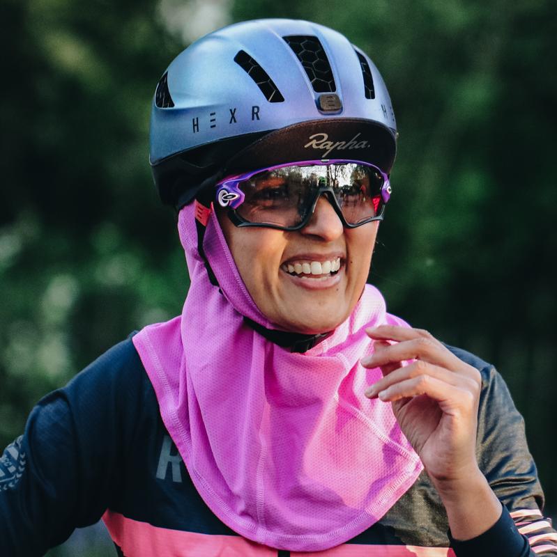 A woman in cycling gear is smiling at the camera. She is wearing a blue cycling helmet with a Rapha cap underneath. She has on cycling sunglasses, a stripey Rapha cycling jersey and a bright pink headscarf