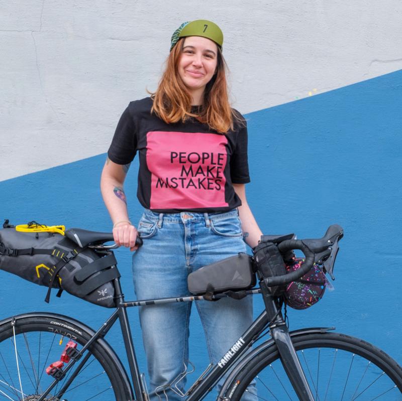 Adna stands smiling with her black bike. She is wearing a cap, black and pink slogan t-shirt and jeans. 