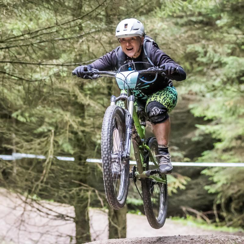Emma is smiling as her bike does a jump on a mountain biking trail. She is wearing a helmet, dark coloured top and green leggings.