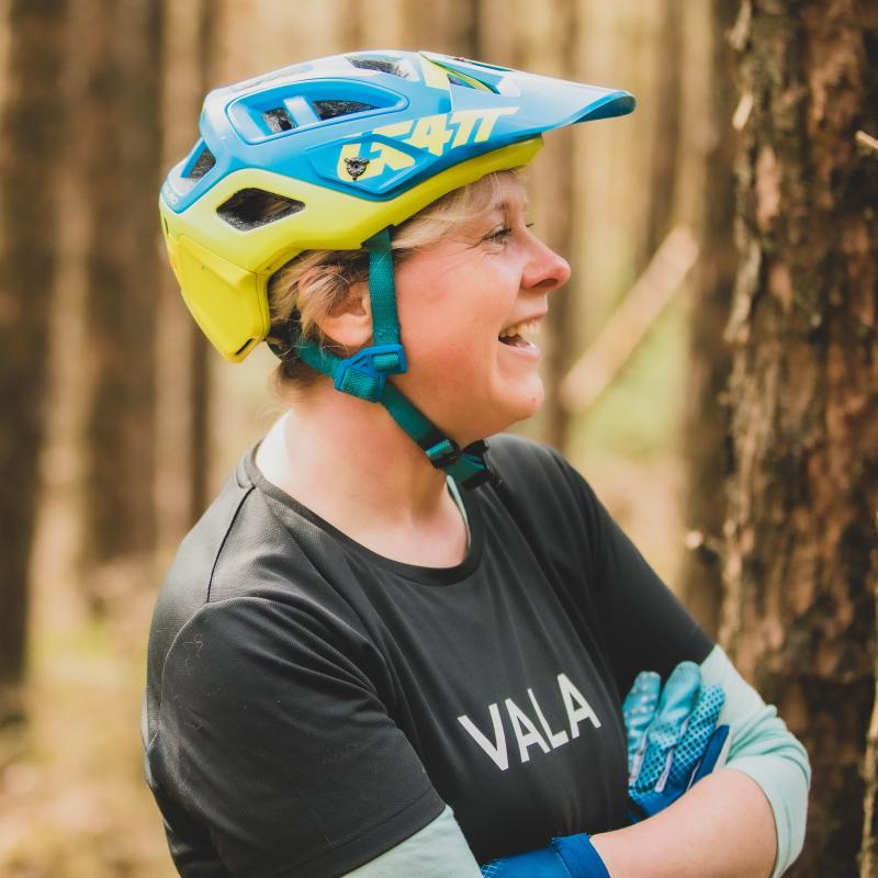Emma smiles with her arms folded. She is wearing a blue and yellow helmet, black top and blue gloves. There are trees in the background.