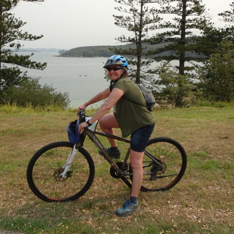 Helen smiling on her bike. She wears a blue helmet, green top and dark shorts. There is a lake behind her.