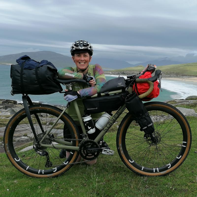 Iona kneels behind her green bike which is set up with bikepacking bags. She wears a helmet, green jersey, gloves and black shorts. A beach is in the background. 