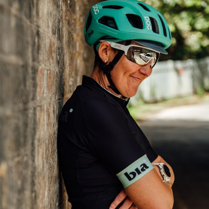 Jenni stands leaning against a brick wall with her arms folded. She wears a green helmet, sunglasses and black jersey. 