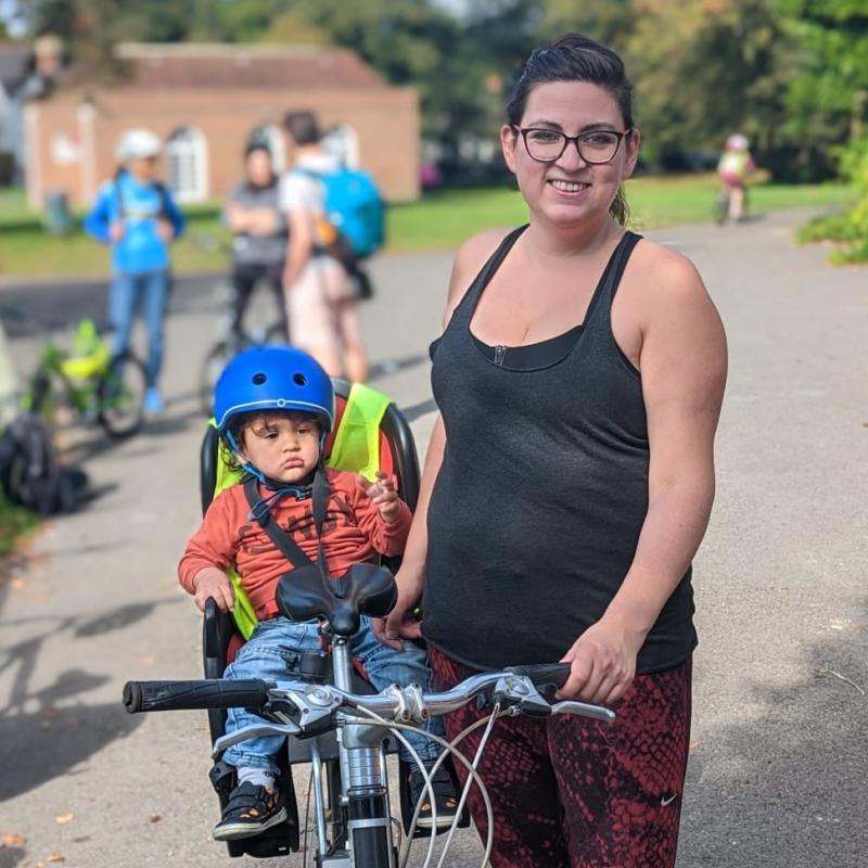 Kat stands to the right of her bike in a park. A small child is in a child’s seat at the back of the bike. Kat is wearing glasses, a black top and red patterned leggings. 