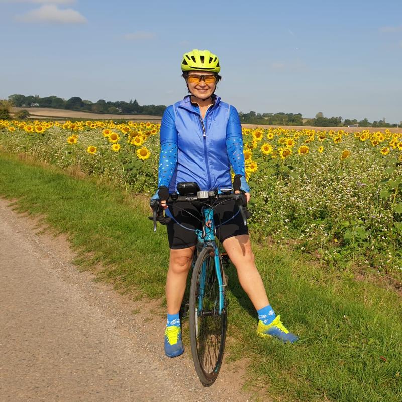 Marina stands next to a sunflower field with her blue bike. She wears sunglasses, a yellow helmet, blue jacket and black shorts. 