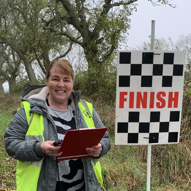 Sharon smiles to camera, holding a clipboard and stopwatch. She is standing at the side of the road at the finish line. She is wearing a grey winter coat and hi-vis vest.