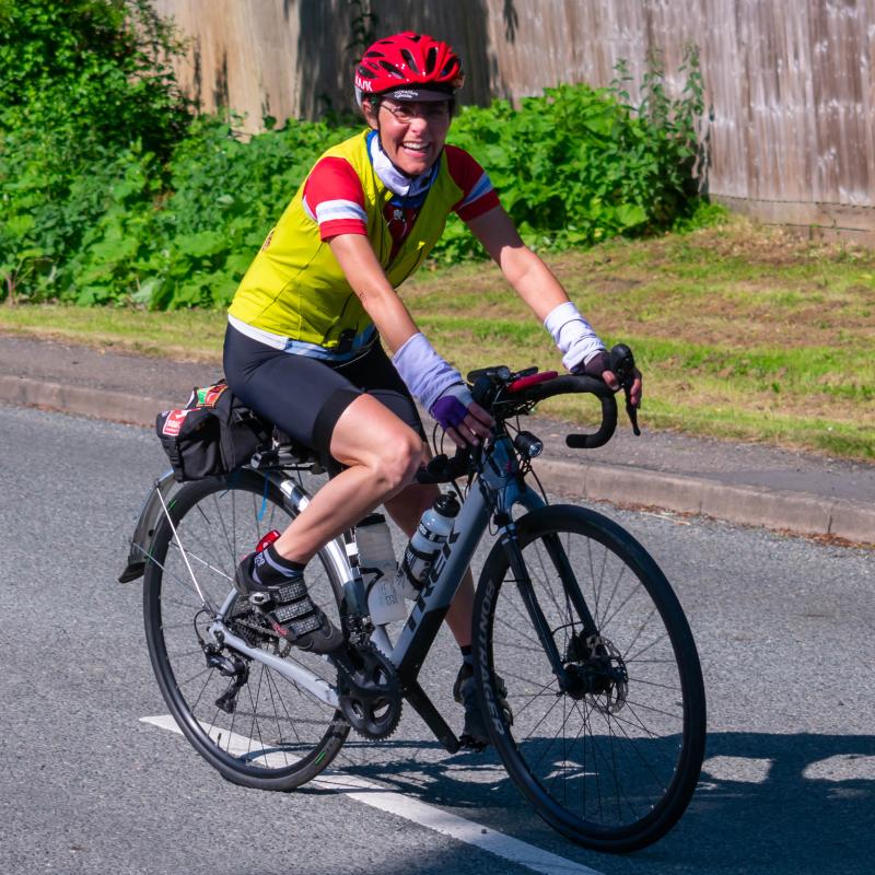 Yasmin smiling as she rides a white Trek bike on road. She is wearing a red helmet, yellow gilet over a red jersey and black cycling shorts. 