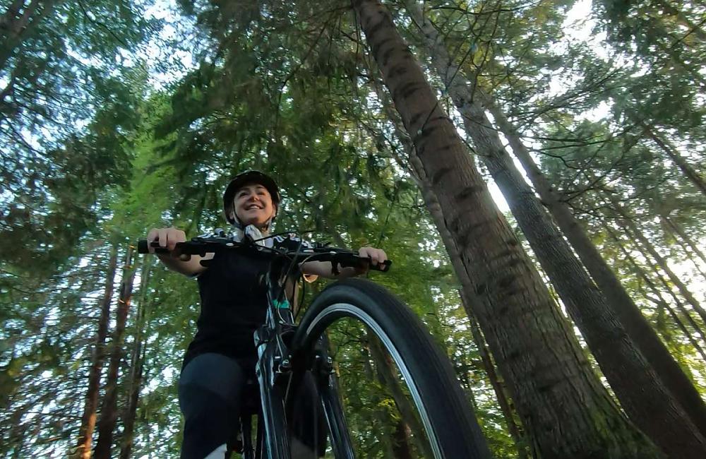 The camera looks up at a woman in dark clothing rides her bicycle through a woodland area